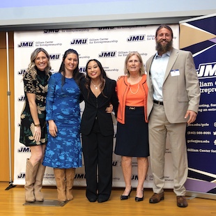 Five adults standing together at a JMU-branded event backdrop, posing for a group photo.