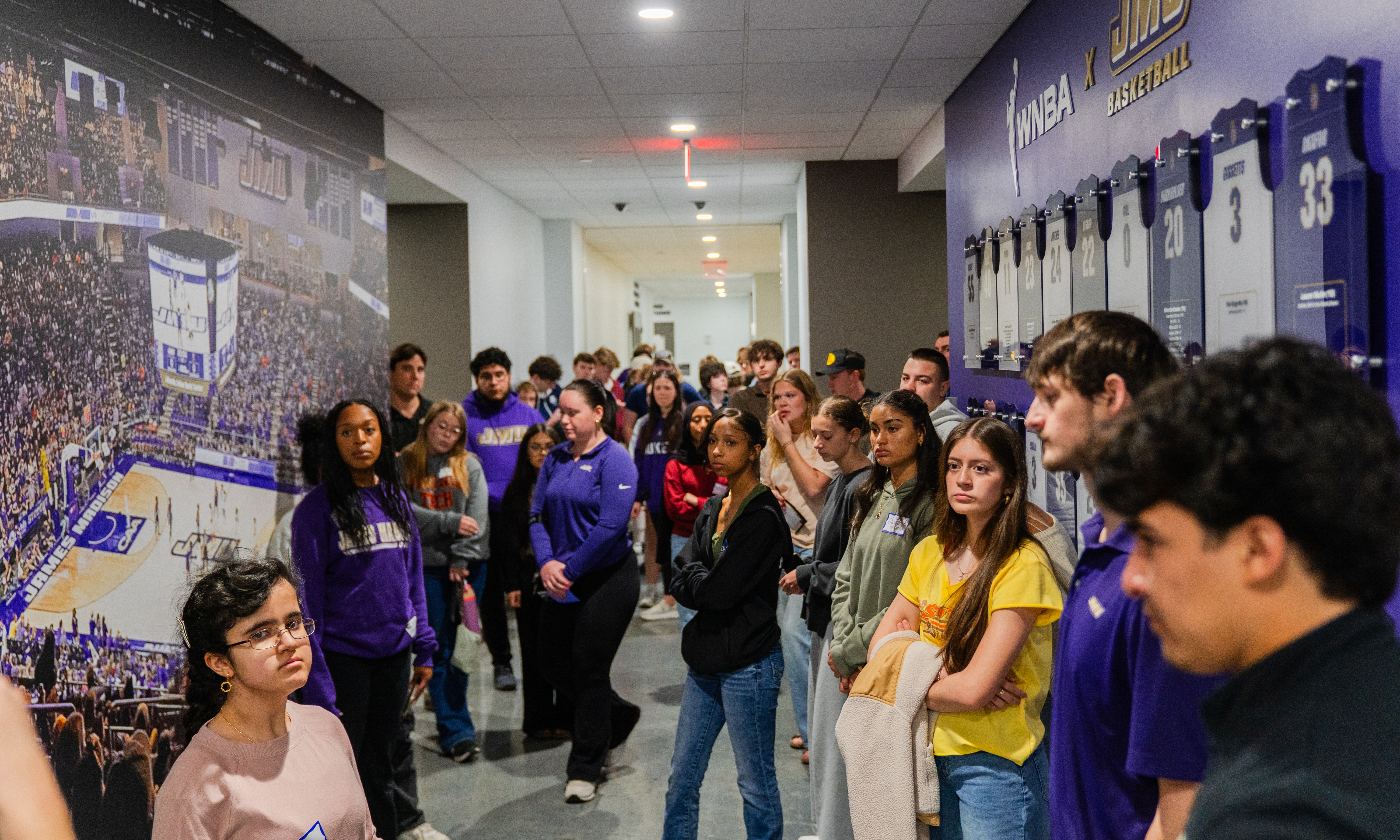 Students gather in a sports arena hallway, looking at a large crowd mural and a wall of basketball jerseys.