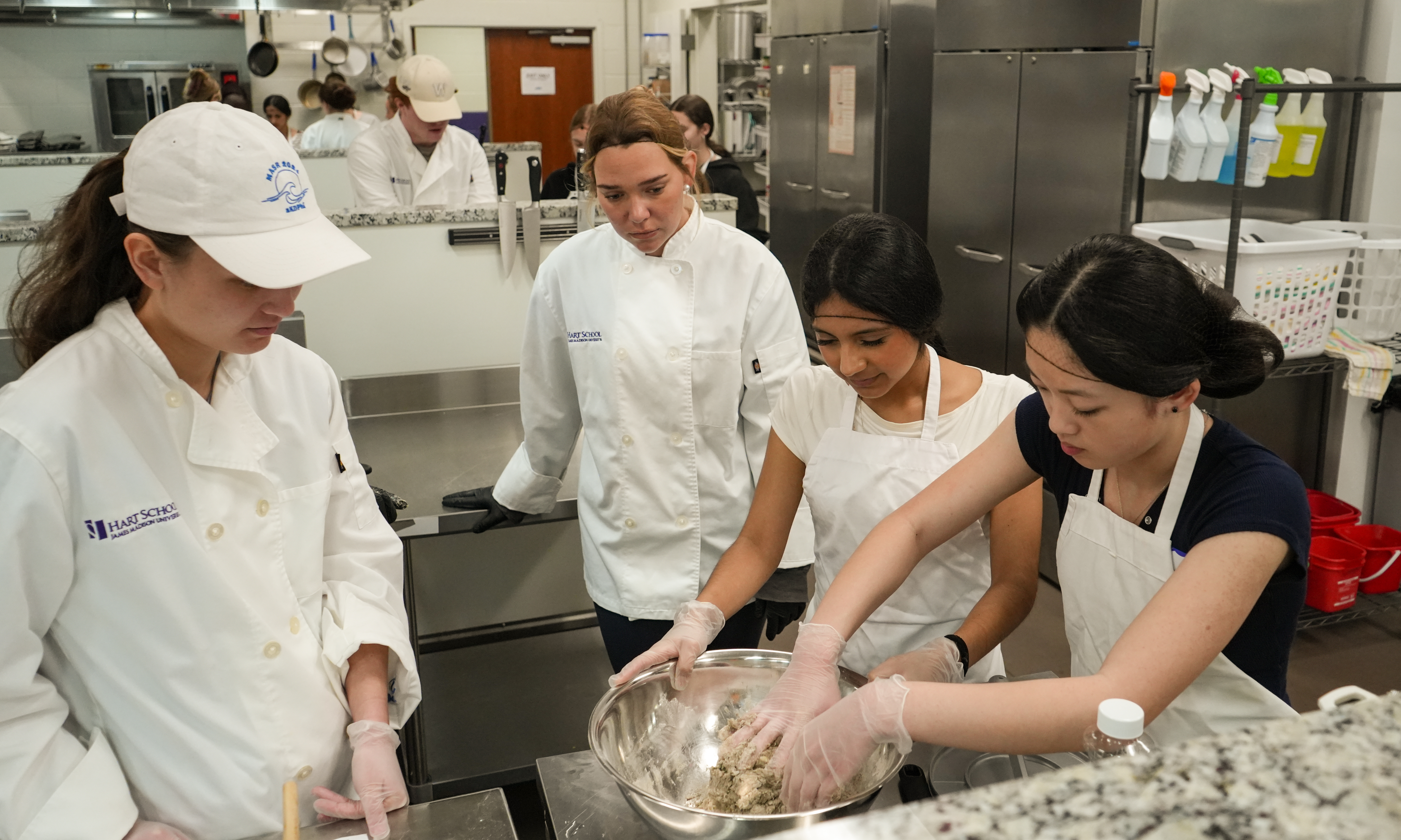 Culinary students in a commercial kitchen mix ingredients in a large metal bowl while an instructor watches.