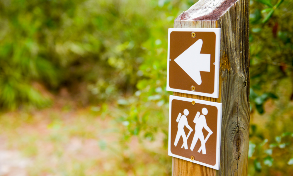 Image of signpost with brown arrow and hiker markers in foreground. Dirt hiking trail with tree foliage out of focus in background. 
