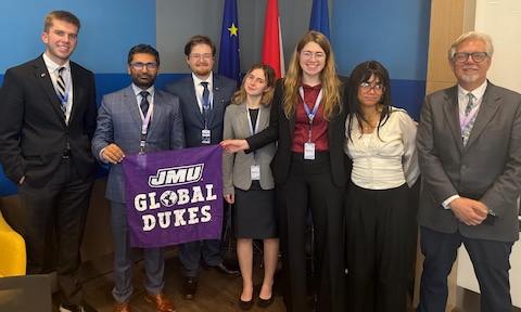 Photo of 7 JMU sudents and staff holding a flag that says 