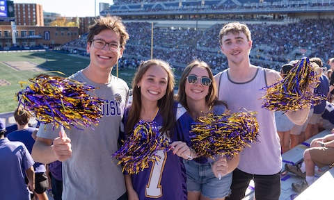 Students cheering at a JMU football game surrounded by purple and gold streamers flying through the air.
