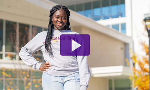 Portrait of a student standing outside a residence hall. Video play button.