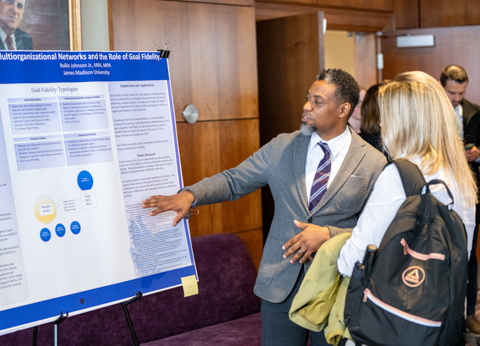 A speaker presenting at a conference in Harrisonburg, VA, with an audience engaged in the background.