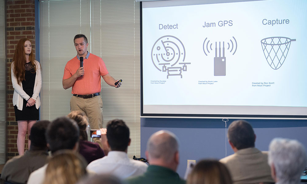 Students discuss slide being shown on a screen. The slide has icons showing the system's capabilities to detect, jam GPS and capture.