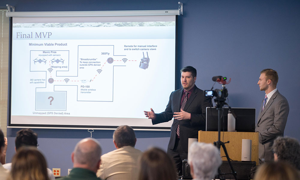 Students discuss slide on screen showing an outline of their system of using drones to perform reconnaissance.