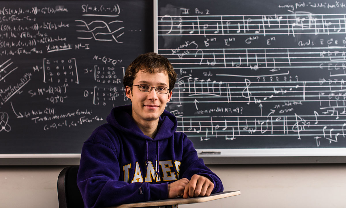 Ryan Stees sits at a student desk in a classroom looking at the camera, wearing a purple JMU sweatshirt. In the background is a blackboard with music written on one side and math formulas on the other.