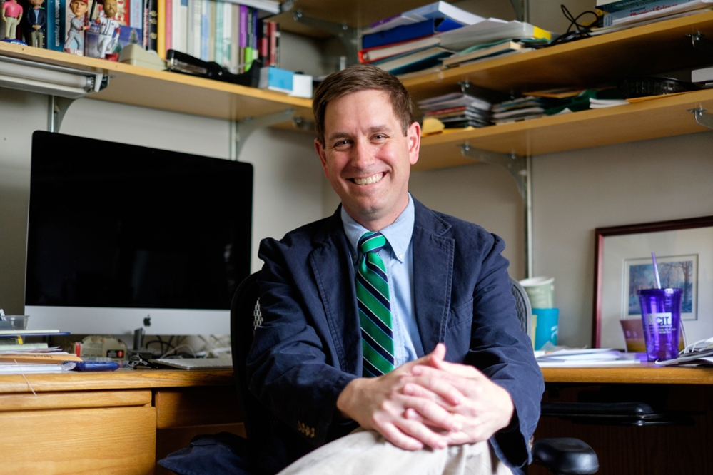 Dr. Chris Hughes seated, facing the camera with his desk and book shelves behind him.