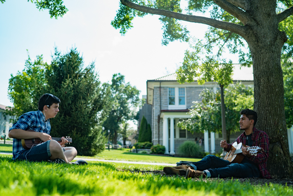 Two students sitting on the Quad playing guitars. 