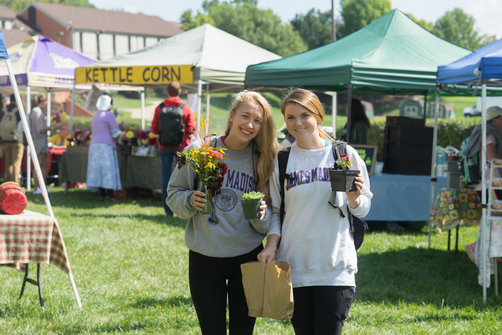 Two girls smiling outside of an outdoor farmers market holding flowers and potted plants