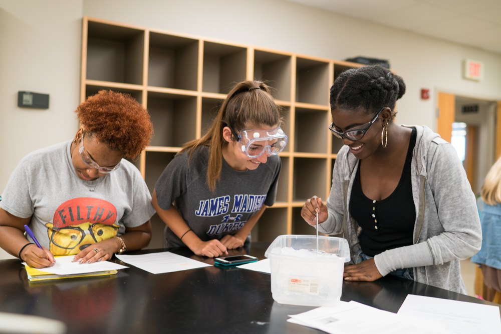 Three students wearing goggles at a lab table, smiling, while studying liquids 