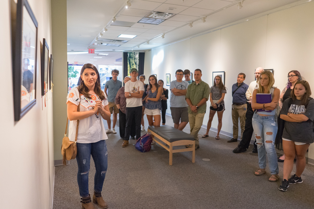 a student standing in front of a group of peers, talking about artwork on the wall. 