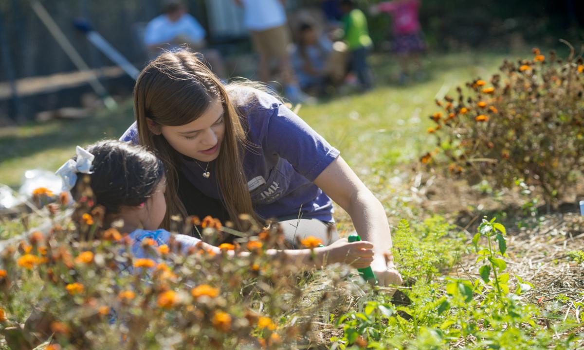 JMU student sitting beside an elementary school student, helping her dig with a hand trowel.
