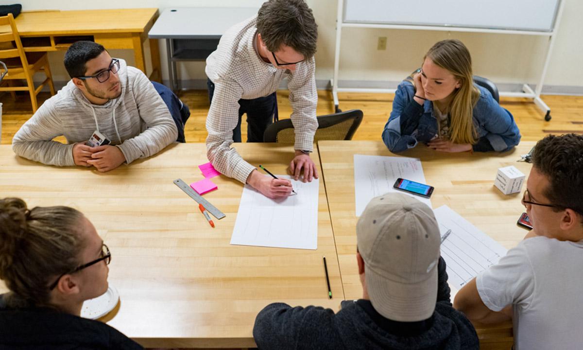 Overhead shot of students working at a table, the middle student is drawing on a piece of paper 