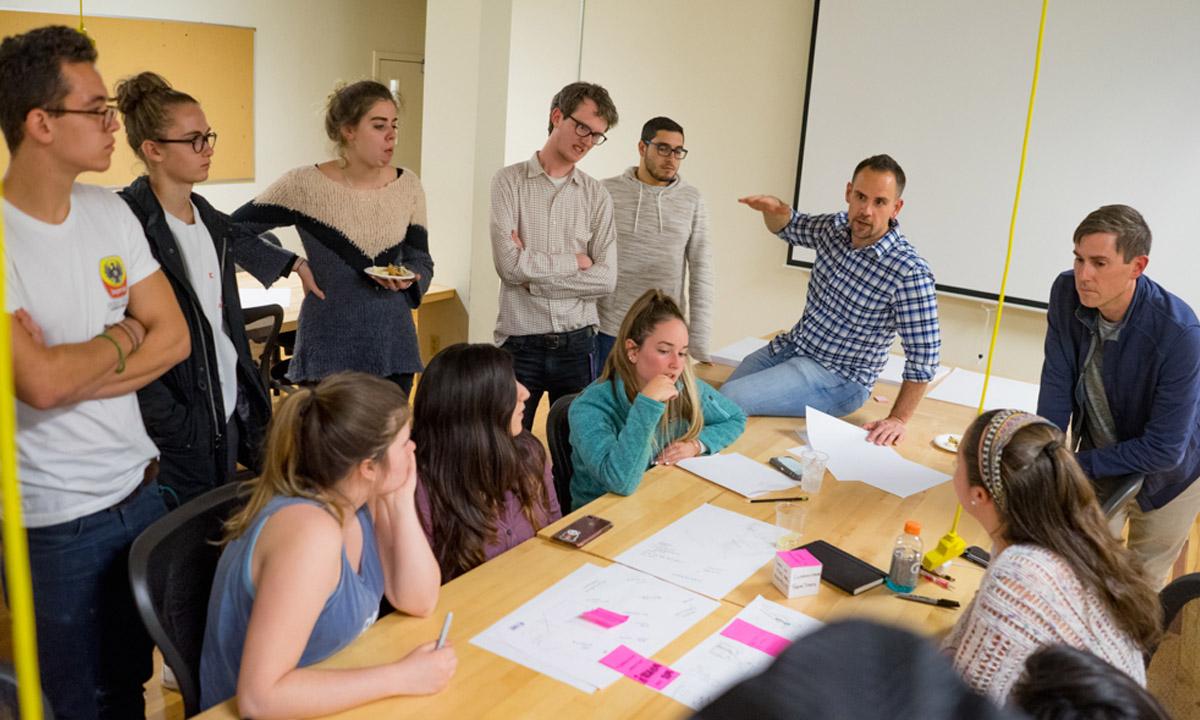 A Professor dynamically talking to students standing around a table.