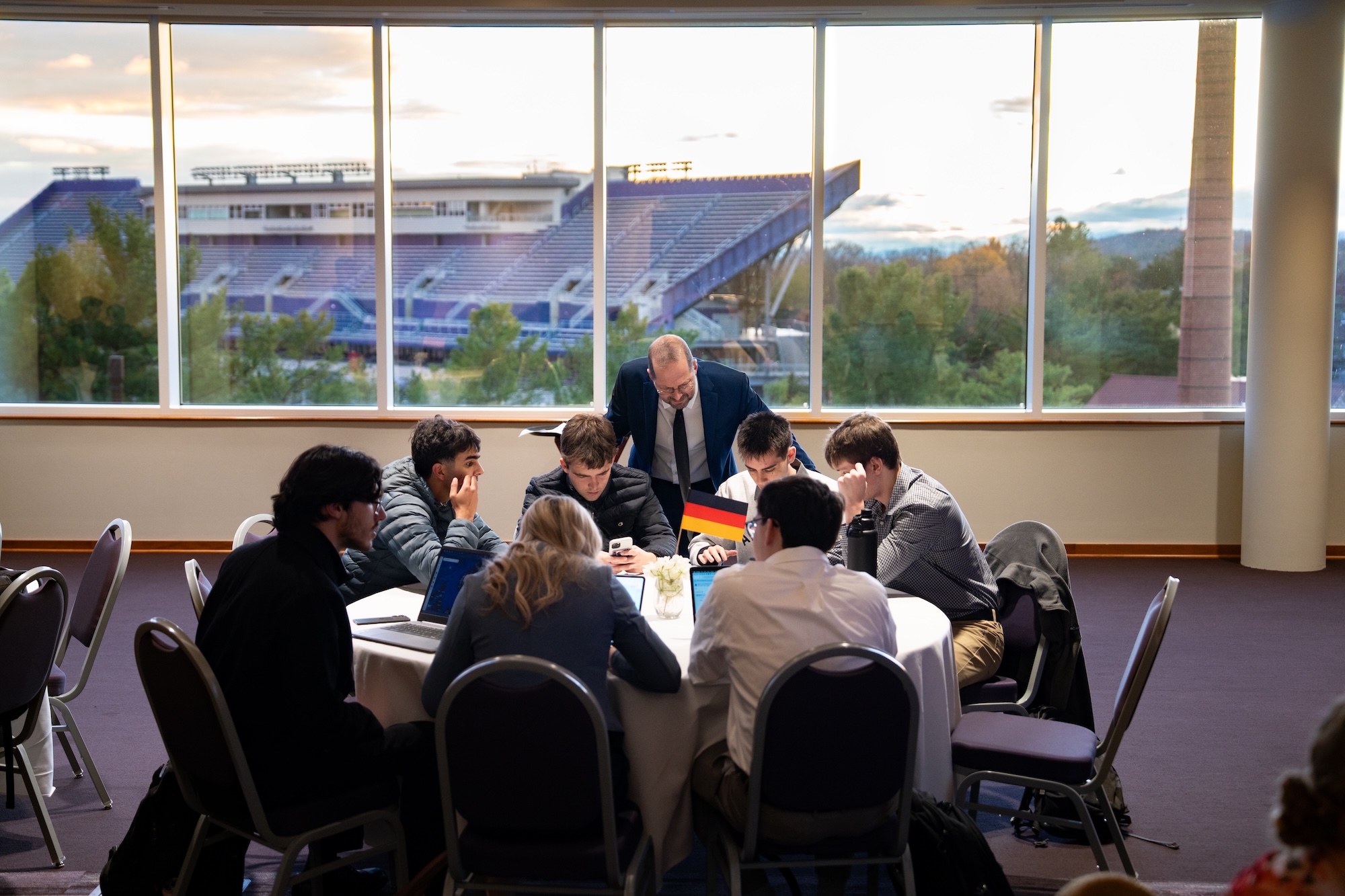Group of students collaborating at a round table, with a professor guiding them, and a stadium visible through the large windows in the background.