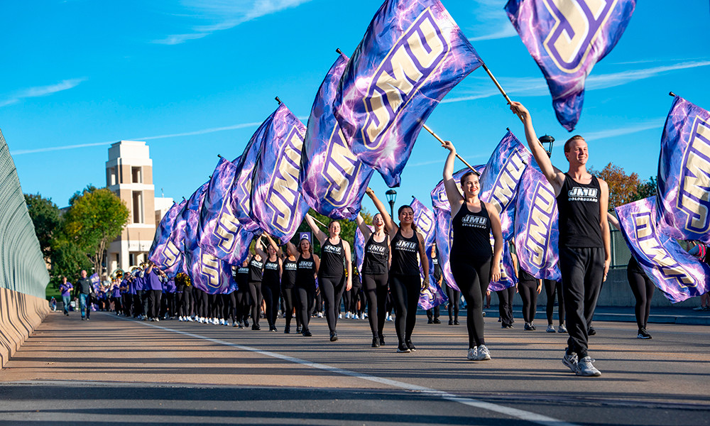 The JMU color guard wave their flags while crossing the Carrier Drive bridge in the Homecoming parade, with King Hall in the distance.