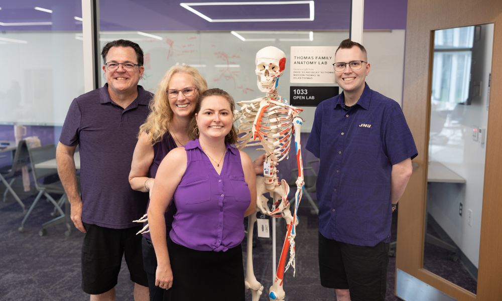 Four people stand smiling beside a labeled human skeleton model in a university anatomy lab hallway.