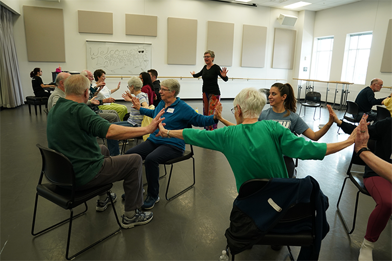 Participants sit in chairs with two rows facing each other. Each participant has their arms outstretched to either side toughing the palms of the participant next to them.