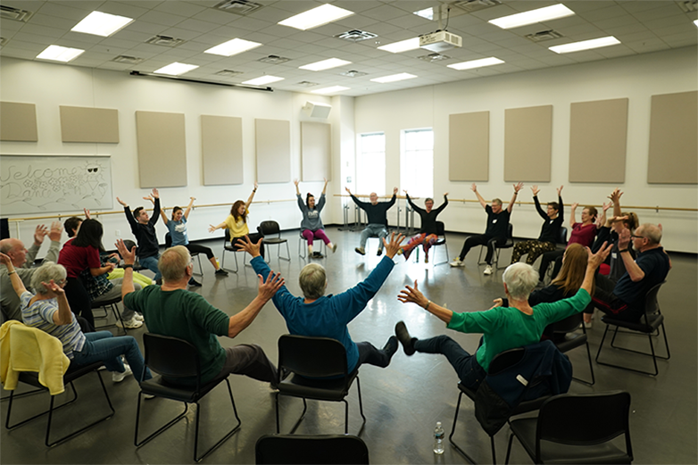 Participants sit in chairs in a circle. They have their arms outreached high and their legs raised from the ground.