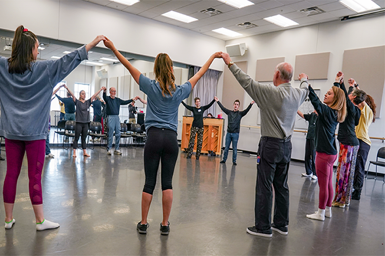 Participants stand in a circle. They have their arms outreached high, holding the hands of the participants next to them.