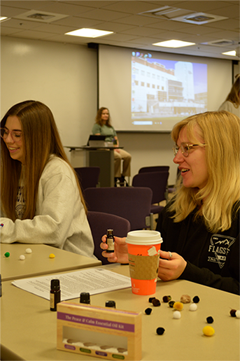 2 women sit at a table with bottles and colored cotton balls, one holds a bottle.