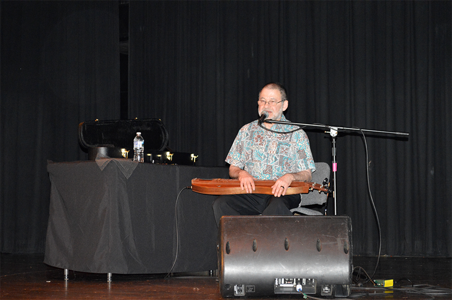 Man sits speaking into a mic with a flat instrument laying across his lap