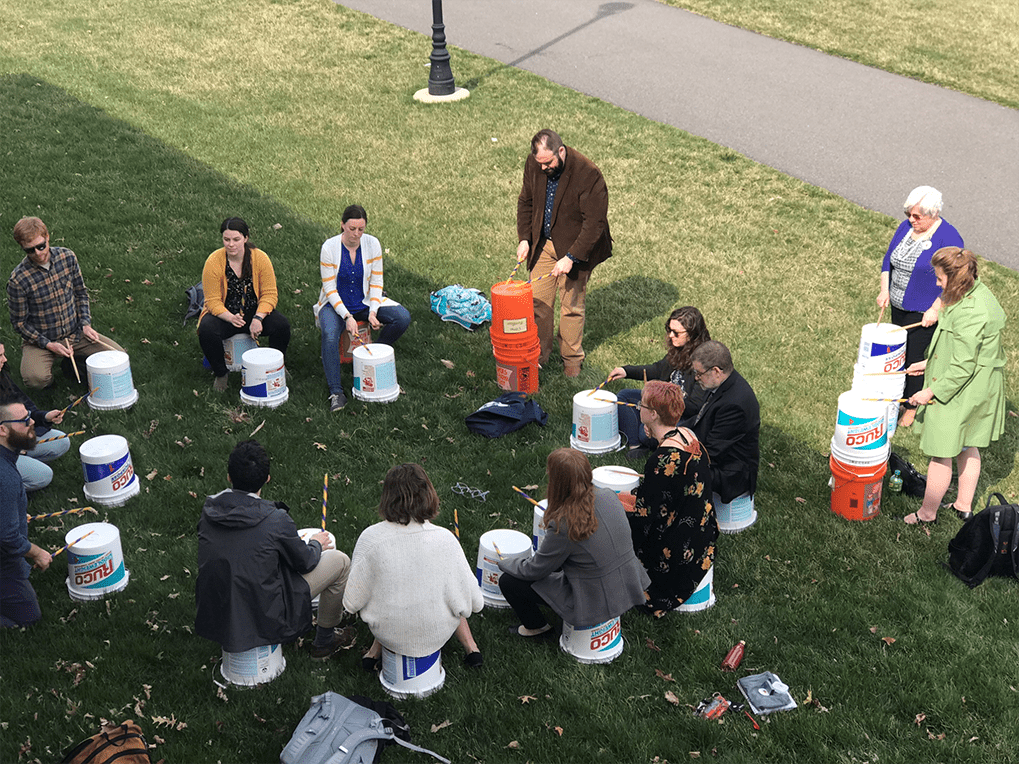 people sit and stand in a circle on grass with buckets in front of them holding drumsticks