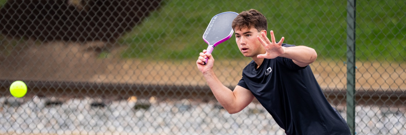 pickleball player prepares to hit a ball near a chain-link fence, holding a racquet up and reaching out with an open hand.