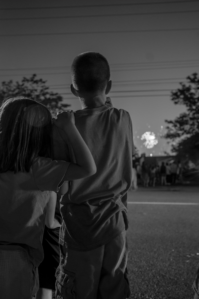 Two children stand, facing away from the viewer as they watch distant fireworks
