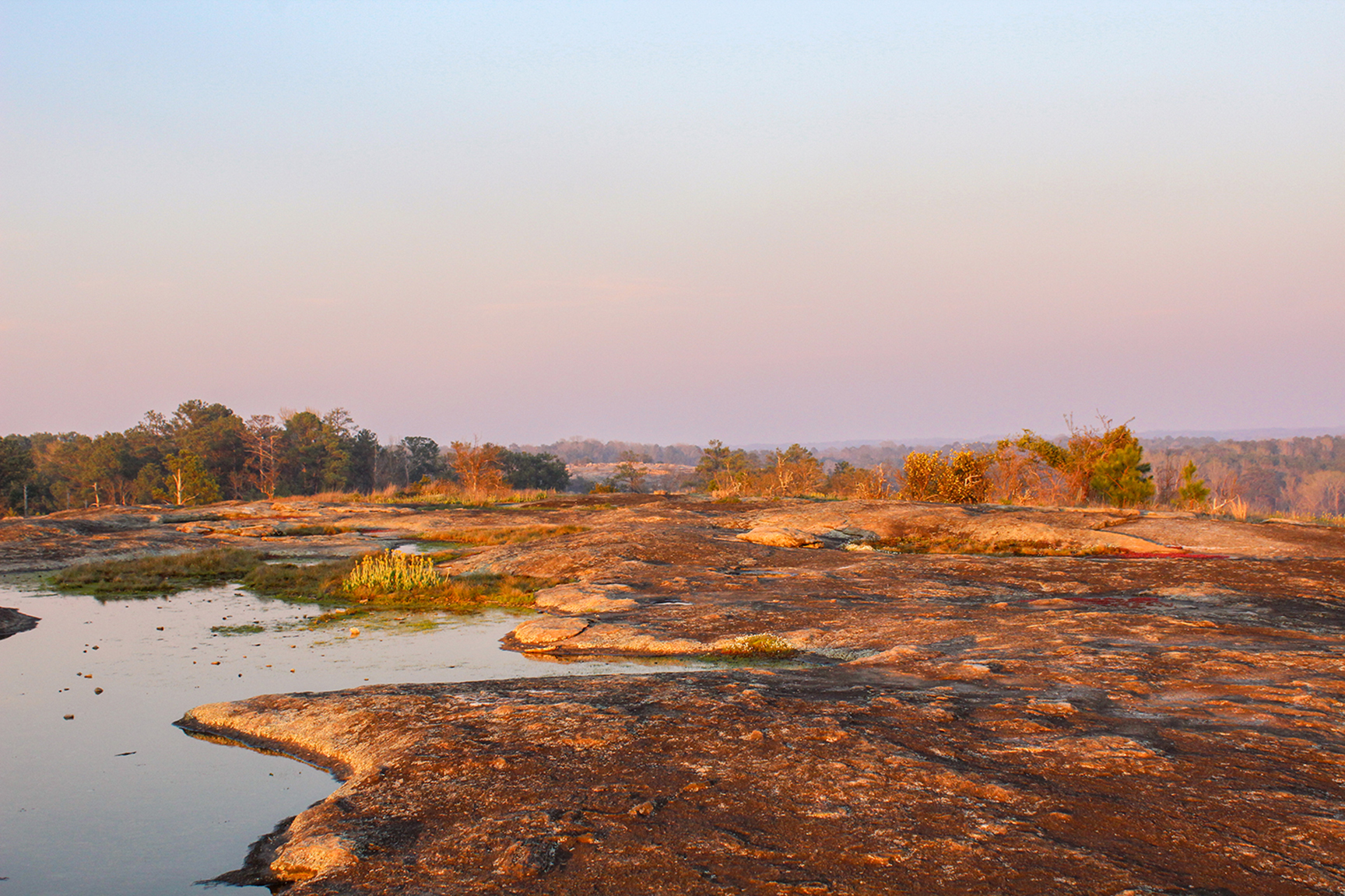 A scenic view of a rocky landscape with a tranquil pool of water and trees in the background during sunset.