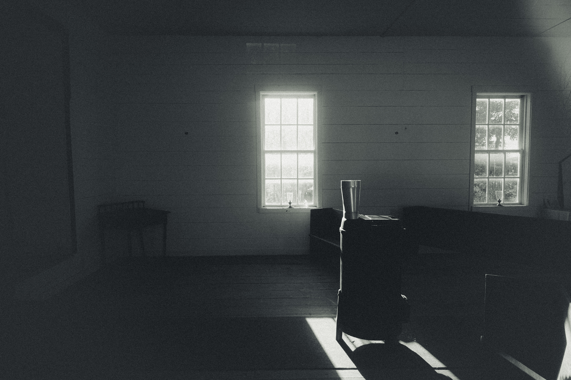 Dimly lit rustic room with sunlight streaming through two windows onto a wooden floor and simple furniture.