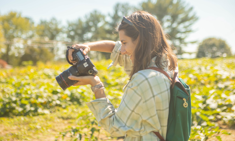 A side profile of a smiling girl in a green, flannel shirt standing in a field, looking at her camera's screen.