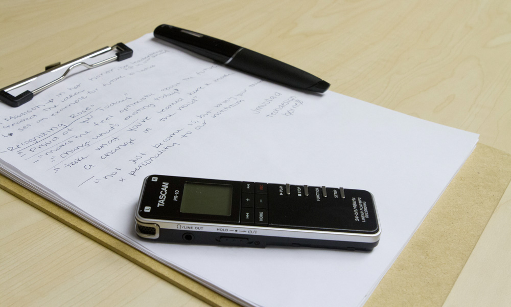 A voice recorder and pen laying on top of a clipboard and paper with notes on written on said paper.