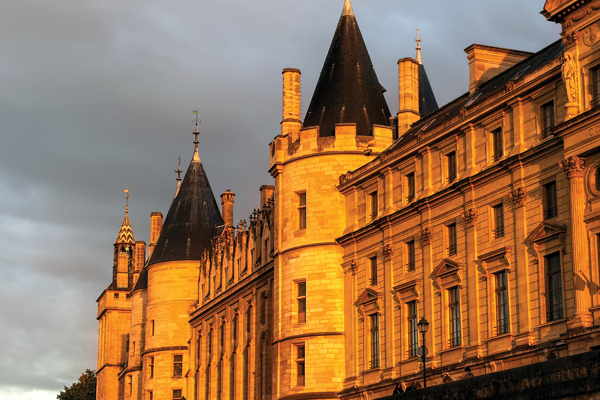 Sunlit historic stone building with ornate windows and tall conical turrets against a cloudy sky.