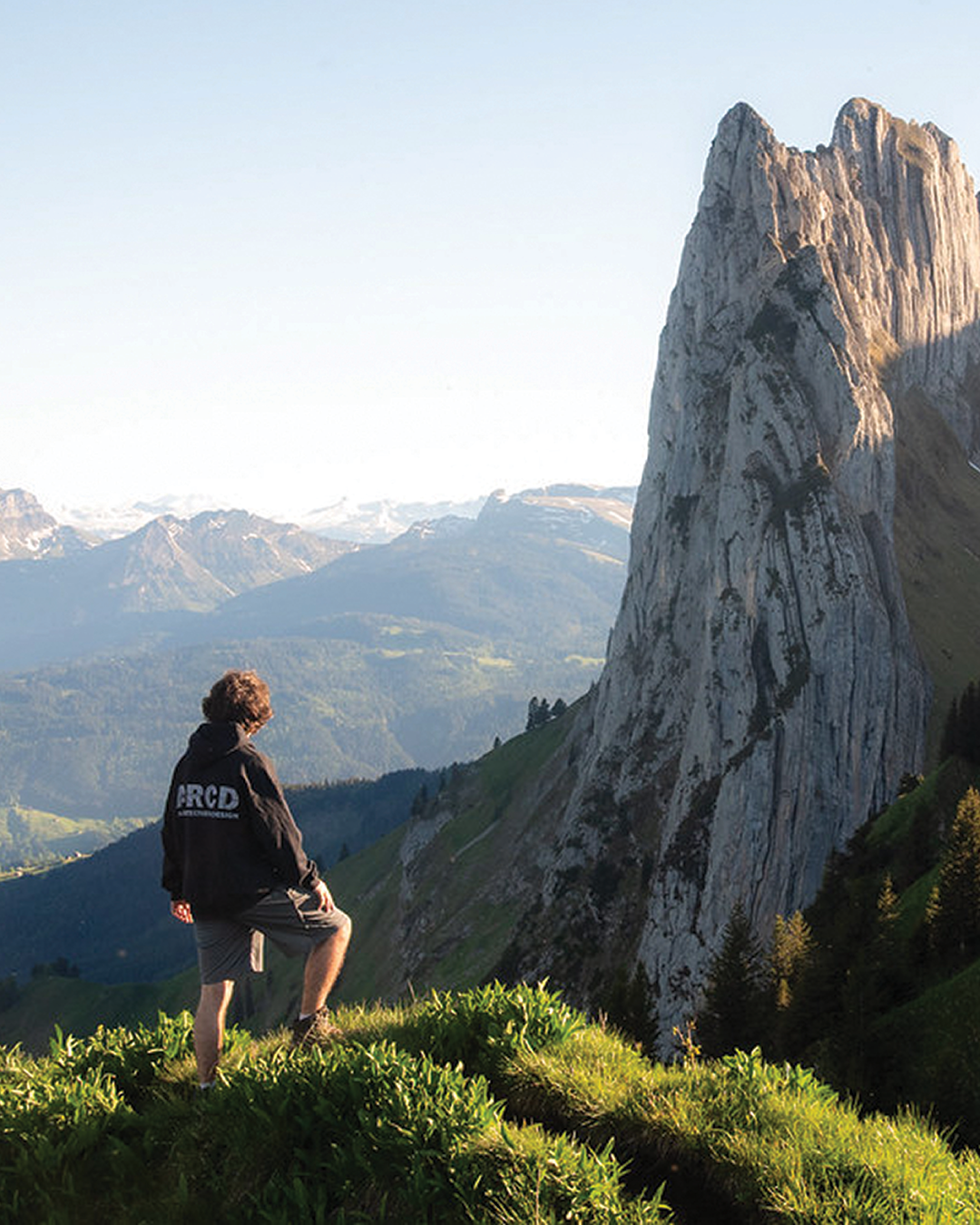 A person standing on a grassy hill, gazing at a towering rock formation against a backdrop of mountains.