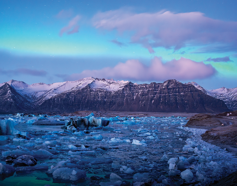 A breathtaking landscape featuring a serene glacial lagoon with floating icebergs, surrounded by snow-capped mountains under an illuminated sky at dusk.