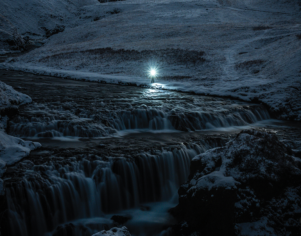 A serene winter landscape featuring a waterfall under moonlight, with a lone figure illuminated in the background.