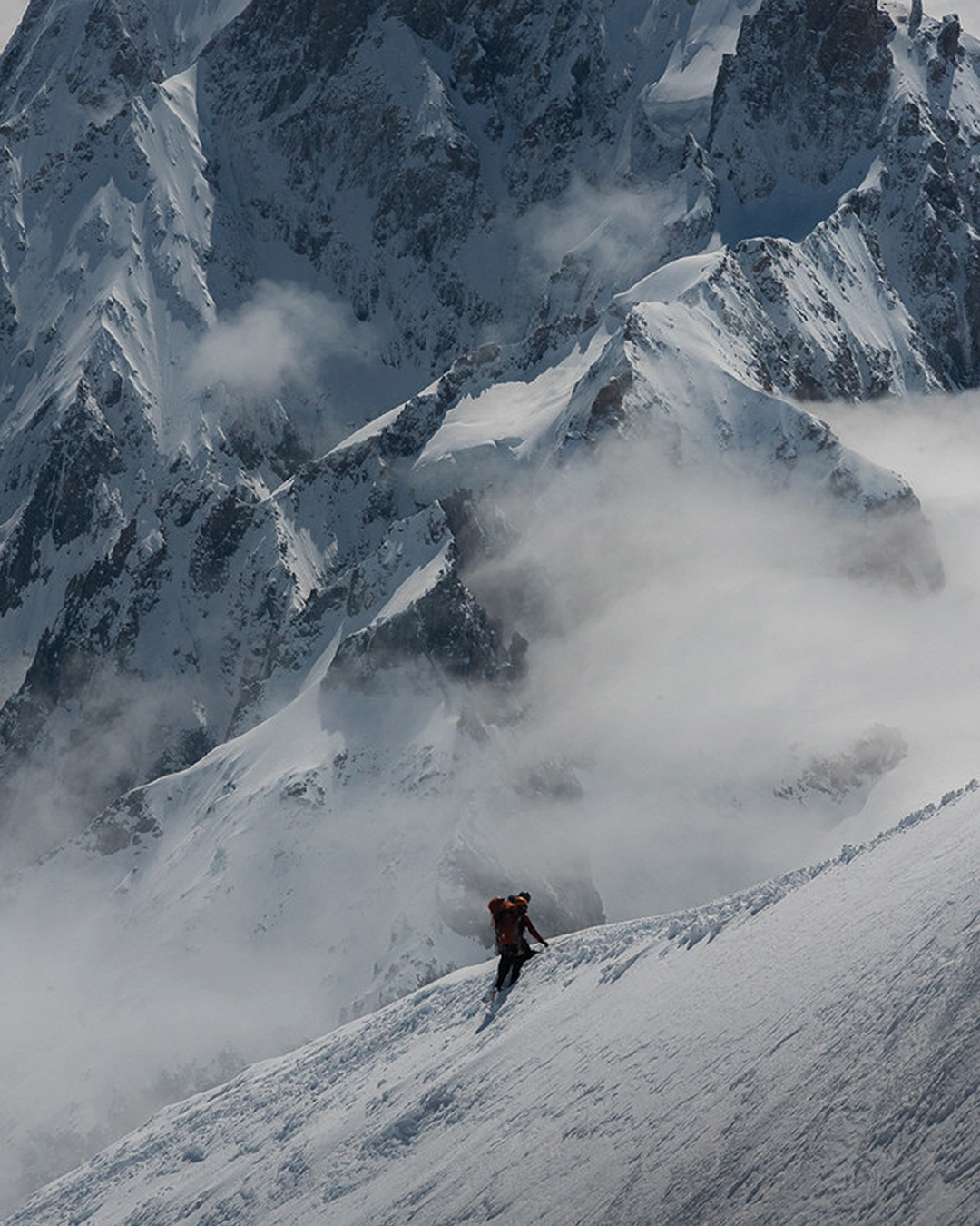 A climber ascends a snowy mountain ridge, surrounded by dramatic, jagged peaks and shrouded in wispy clouds.