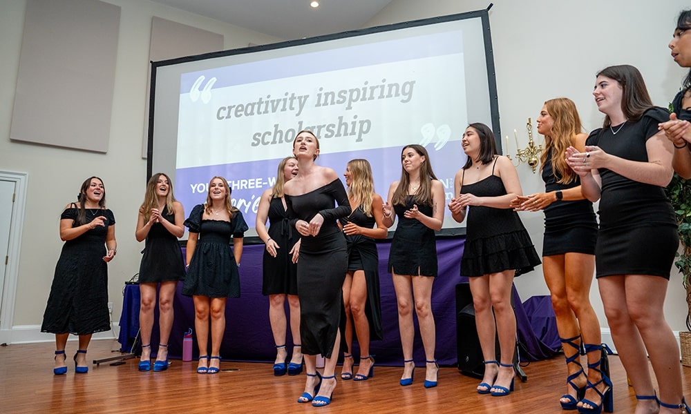 A group of female performers in black dresses and blue heels singing on stage, with a backdrop displaying the words "creativity inspiring scholarship."