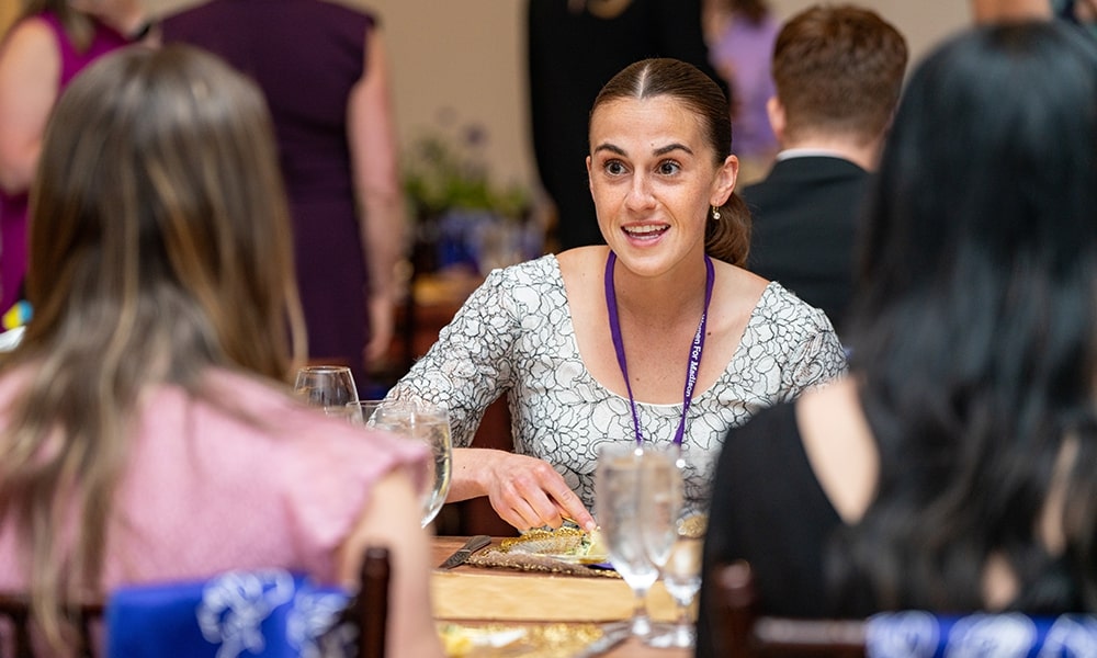 A woman engages in conversation at a social event, seated at a table with food and drinks around her.