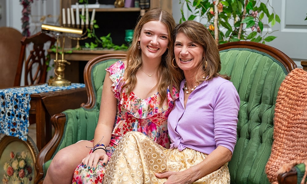 A mother and daughter smile together while sitting on a green couch in a cozy living room setting.