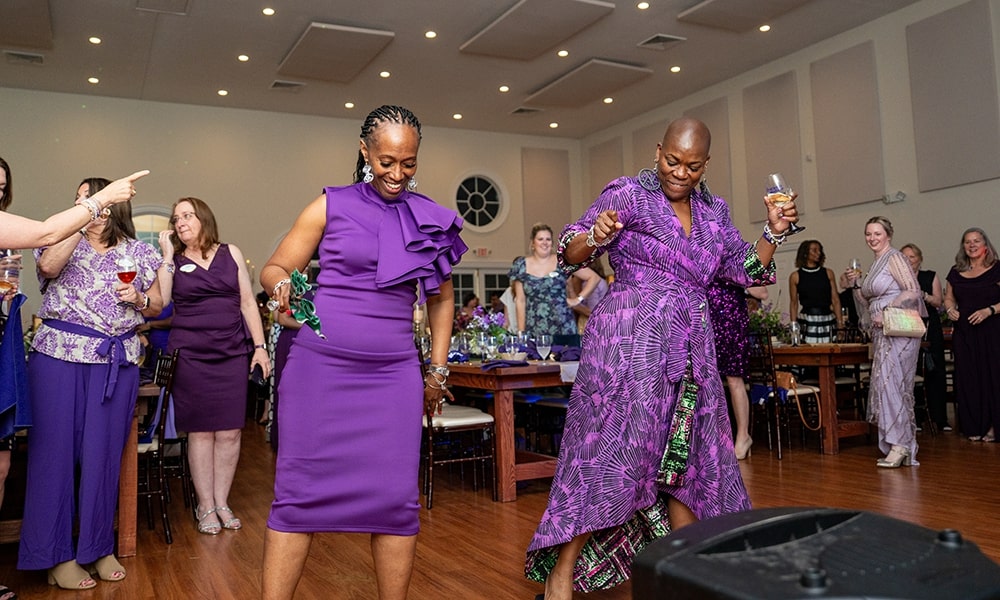 Two women dressed in stylish purple outfits dance joyfully at a lively celebration, surrounded by party guests.