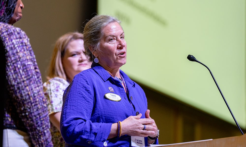 A woman in a blue shirt speaks at a podium, with two others standing behind her.