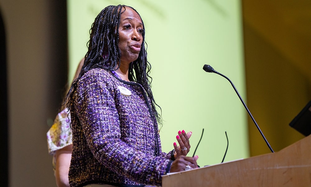 A woman with long braided hair speaks at a podium during a presentation.