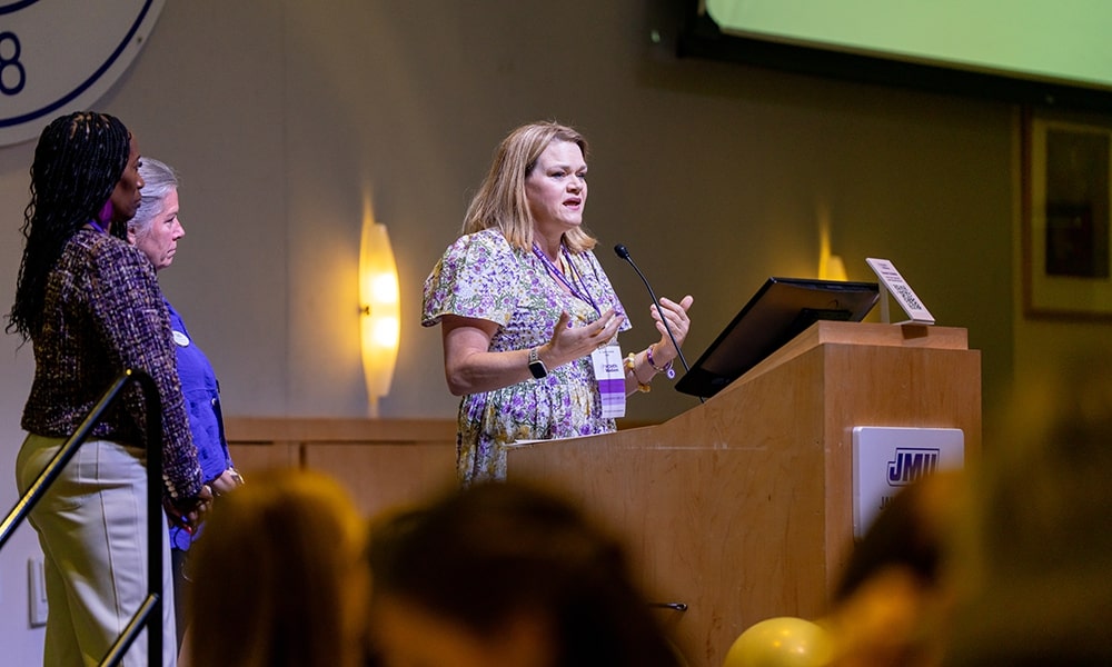 A woman speaks at a podium while two other women listen attentively during a conference event.