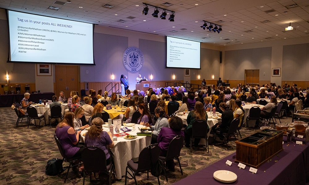A conference event featuring a large audience seated at round tables, with a speaker presenting on stage and social media hashtags displayed on a screen.
