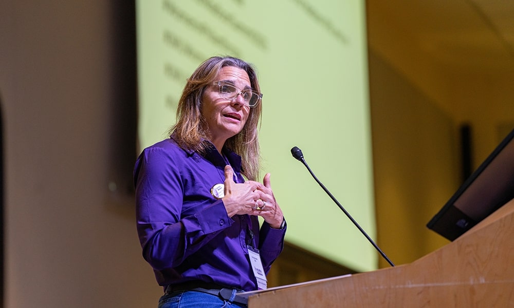 A speaker presenting at a conference, gesturing towards the audience from behind a podium.