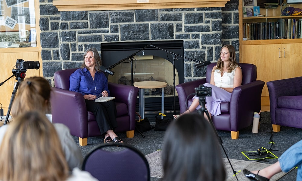 Two women seated in armchairs during a discussion or interview, with microphones and cameras set up in a cozy indoor setting.