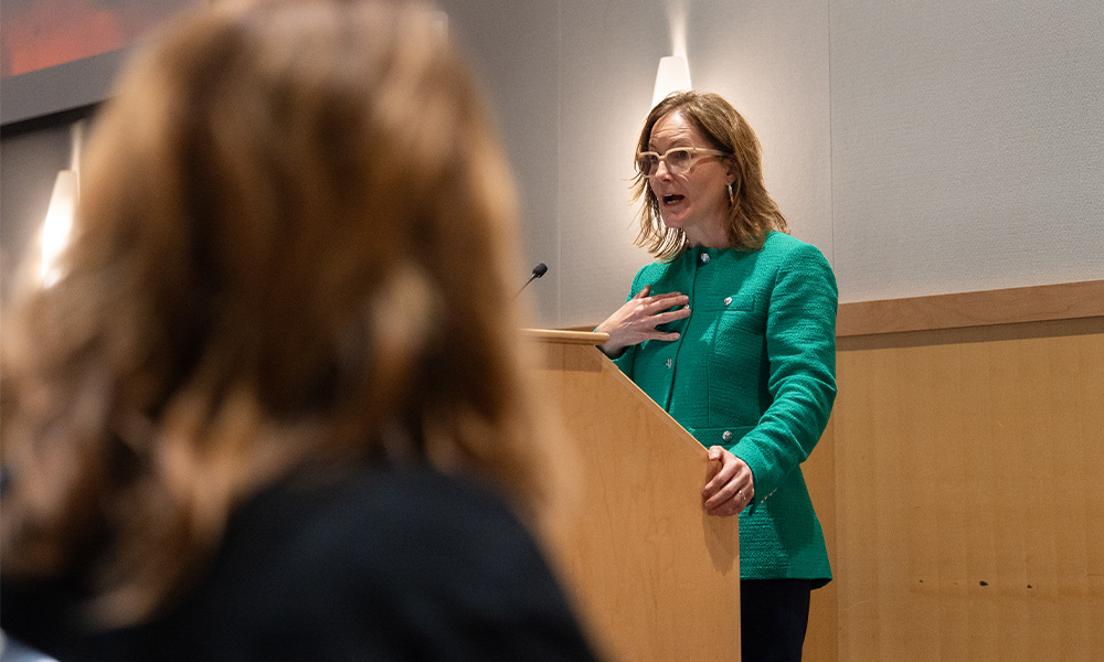 A woman in a green blazer passionately speaks at a podium, engaging with an audience.